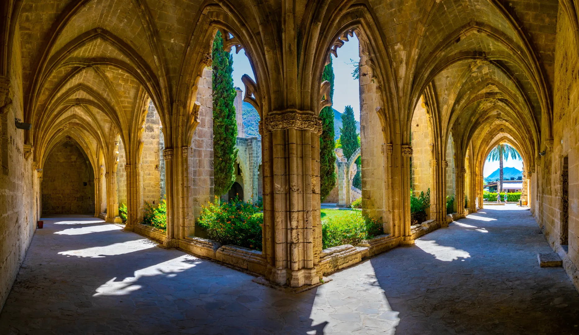 A wide-angle view of the Gothic cloisters at Bellapais Abbey in the village of Bellapais, North Cyprus. The image showcases the intricate stone-ribbed vaulted ceilings and pointed arches of the 13th-century monastery. Sunlight streams through the arches into the cool, shaded walkway, overlooking a lush central courtyard with tall cypress trees and flowering gardens, with the Kyrenia Mountains visible in the distance.