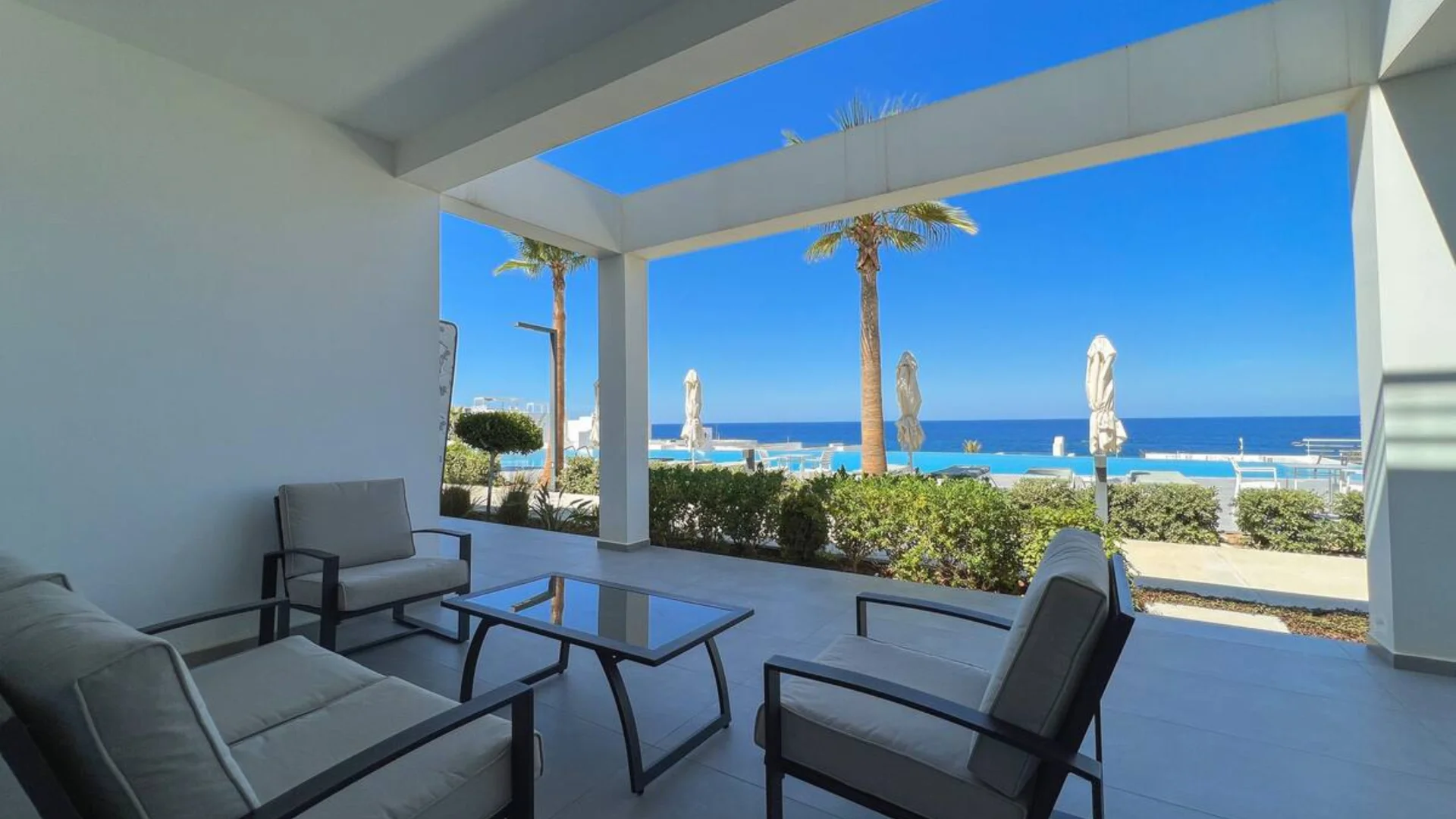 A view from a modern, shaded outdoor lounge area at a luxury residential complex in Esentepe, North Cyprus. The terrace features grey cushioned seating and a glass coffee table, looking out over a blue infinity pool, palm trees, and the Mediterranean Sea under a bright, clear sky.