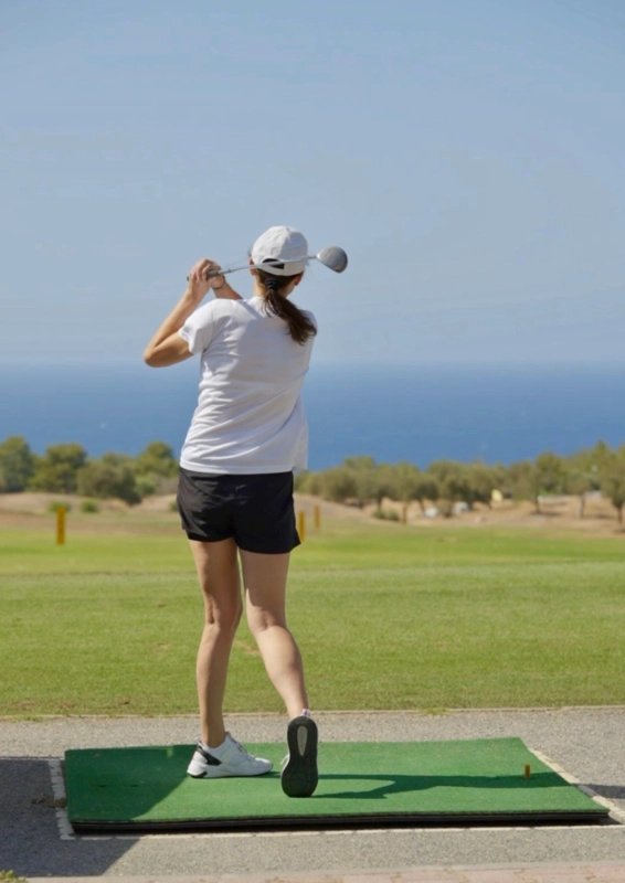 A rear view of a female golfer in mid-swing on a practice mat, overlooking a lush green course. She is wearing a white cap, a white t-shirt, and black shorts, with the deep blue Mediterranean Sea and a clear sky visible in the distance at Korineum Golf & Beach Resort in Esentepe North Cyprus.