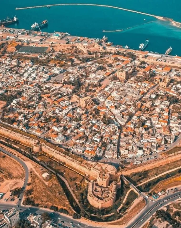 An aerial panoramic view of the historic Walled City of Famagusta (Gazimağusa), North Cyprus. The image showcases the massive Venetian fortification walls and the Martinengo Bastion in the foreground, with the dense medieval city grid, the Gothic Lala Mustafa Pasha Mosque, and the busy commercial Famagusta Port leading into the Mediterranean Sea.