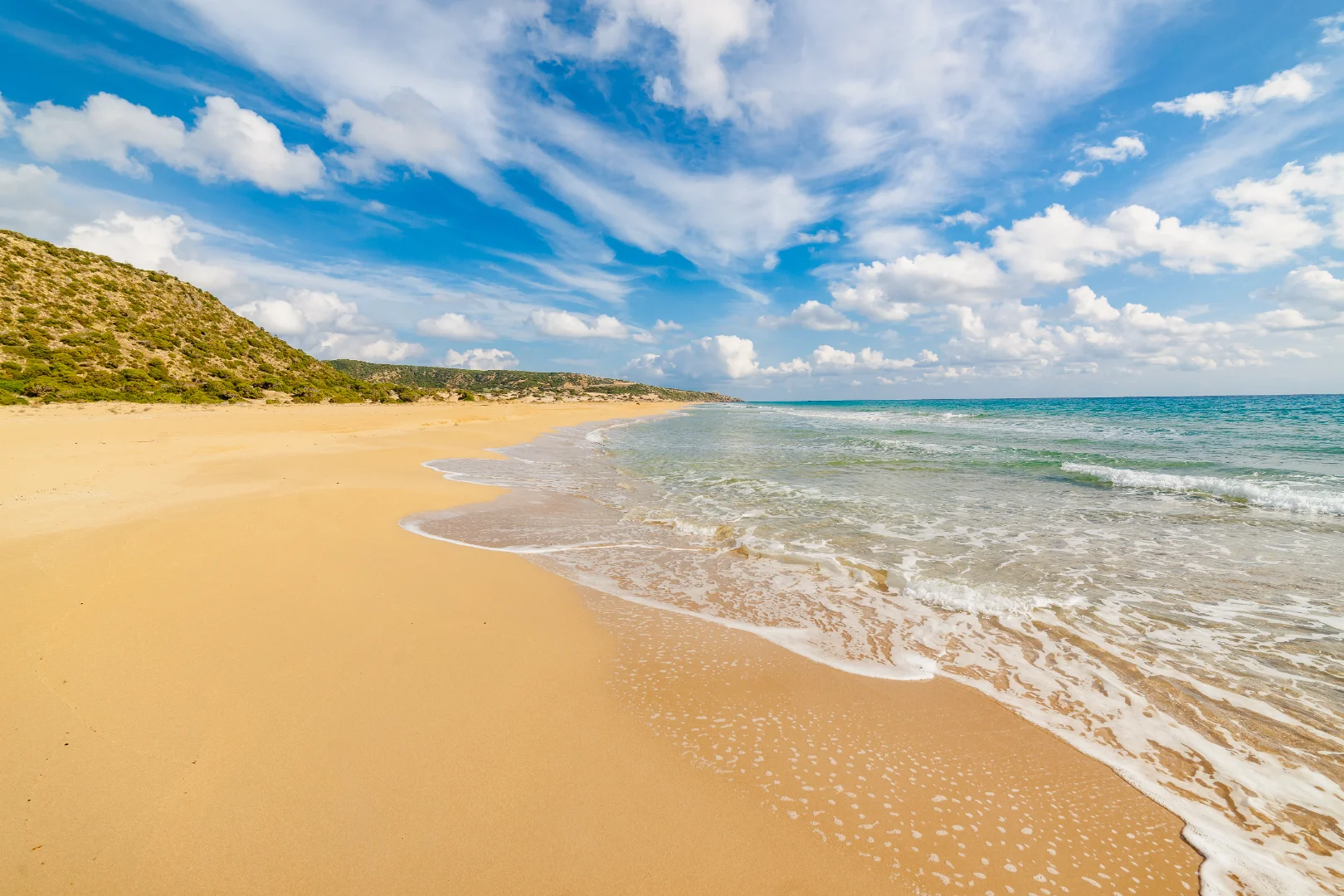 A wide-angle panoramic view of Golden Beach (Nangomi Bay) on the Karpaz Peninsula, North Cyprus. The image shows a vast stretch of untouched, golden sand meeting the gentle waves of the turquoise Mediterranean Sea. Rolling green dunes and low hills line the shore under a bright blue sky filled with wispy white clouds, capturing the wild, natural beauty of the region.