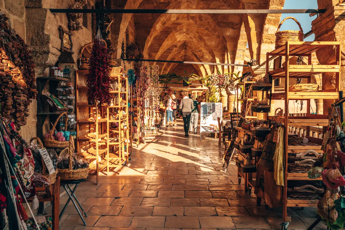 The sun-drenched interior arcade of Büyük Han, a 16th-century Ottoman caravanserai in Nicosia, North Cyprus. The arched stone walkway is lined with rustic wooden shelves and woven baskets displaying colorful Cypriot handicrafts, dried red peppers, and local souvenirs. Visitors stroll through the long, vaulted corridor where warm sunlight creates long shadows on the polished stone tile floor.
