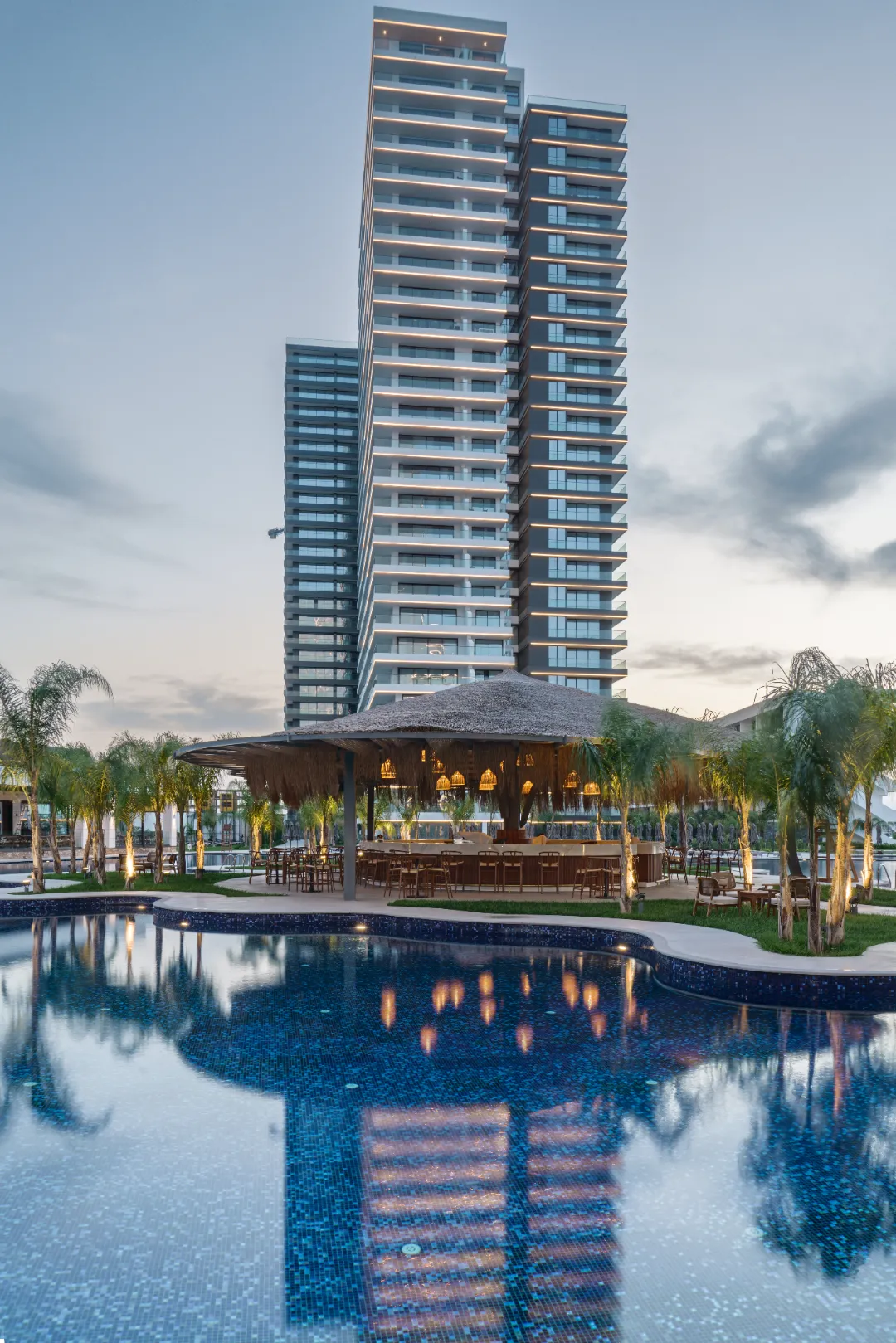 An evening view of the Grand Sapphire Resort & Casino in Iskele Long Beach, North Cyprus, featuring the illuminated Punta Cana pool reflecting the resort's high-rise residential towers. The foreground highlights a modern, thatched-roof pool bar surrounded by palm trees, set against the backdrop of luxury 27 to 31-story apartment blocks under a twilight sky.
