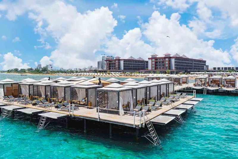 A wide shot of a luxurious Hotel in Iskele Northern Cyprus, featuring a large wooden pier extending over vibrant turquoise waters. The pier is lined with numerous private cabanas with white curtains and modern lounge chairs, while the multi-story hotel buildings and a sandy beach are visible in the background under a bright, cloudy sky.