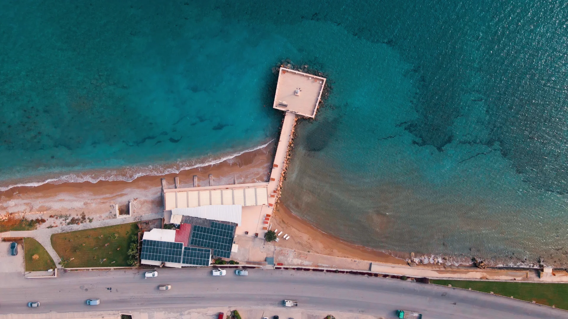 An aerial bird’s-eye view of the Iskele Long Beach coastline in North Cyprus. The image shows a narrow wooden pier extending into crystal-clear turquoise Mediterranean waters, leading to a small square platform at the end. On the shore, a modern building with solar panels sits next to a sandy beach and a coastal road with parked cars, capturing the sustainable and scenic beauty of the region.
