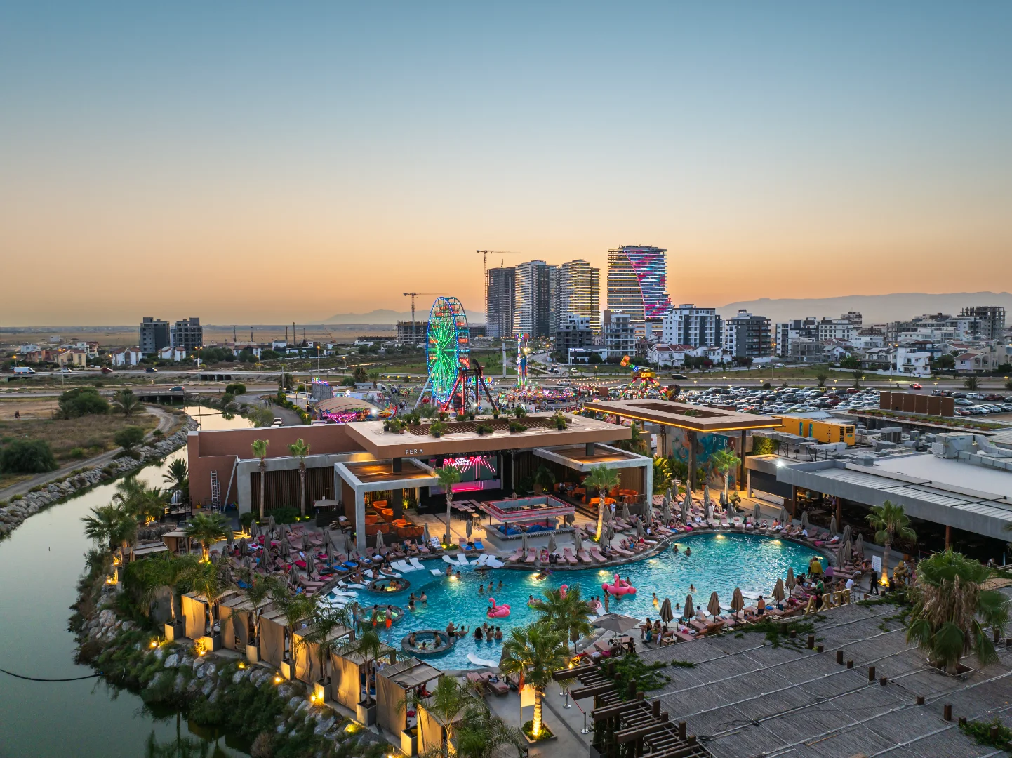 An aerial twilight view of the rapidly growing real estate development in Iskele Long Beach, North Cyprus. The foreground shows the vibrant Pera Mackenzie Beach Club with its illuminated lagoon pool and social areas, while the background features the iconic high-rise towers of Grand Sapphire Resort under construction, a glowing Ferris wheel, and a modern urban skyline against a soft sunset.