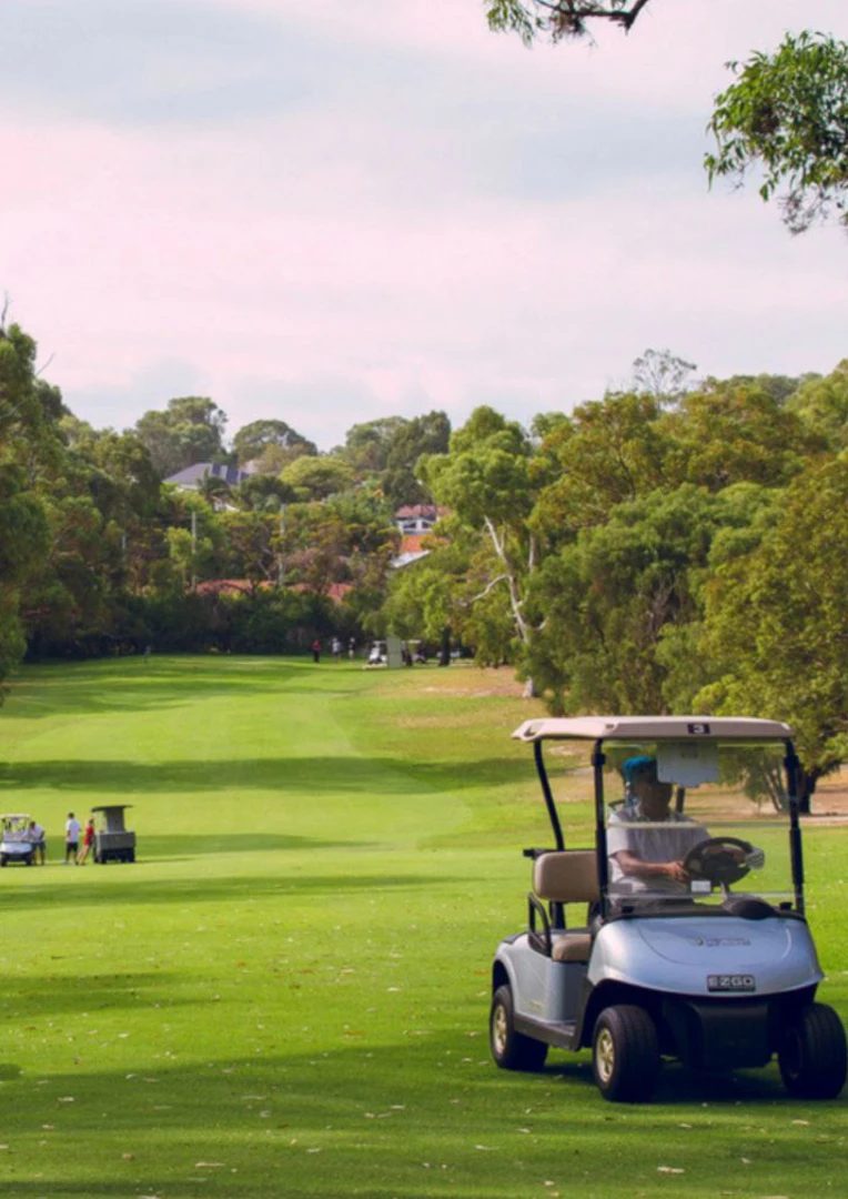 A sunny afternoon at Korineum Golf & Beach Resort in Esentepe, North Cyprus. The image shows a silver EZGO golf cart being driven on a lush green fairway, surrounded by a dense forest of pine and olive trees. In the background, other golfers and carts are visible under a soft, overcast sky, highlighting the premier 18-hole championship golf course and Mediterranean mountain landscape.