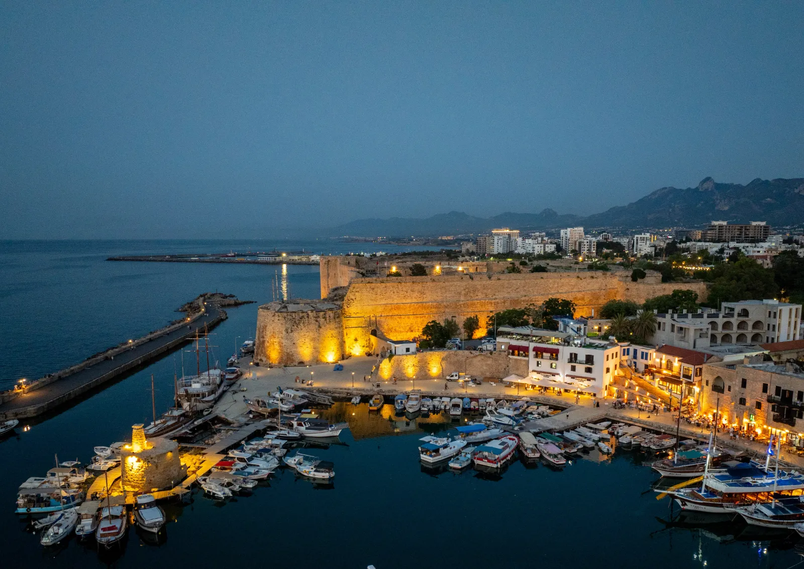 An aerial night view of the historic Kyrenia Harbour and Kyrenia Castle in North Cyprus. The golden-lit ancient fortress walls overlook a semi-circular harbor filled with traditional wooden boats and modern yachts. The waterfront is lined with glowing restaurants and cafes, with the silhouettes of the Kyrenia Mountains and the city skyline visible under a deep twilight sky.