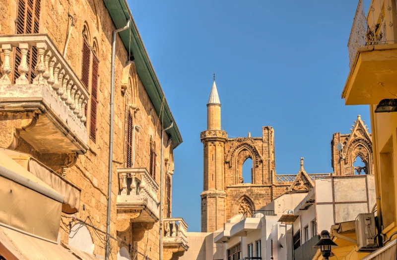A sun-drenched narrow street in the historic Walled City of Famagusta, North Cyprus. The image features traditional stone buildings with arched windows and balustraded balconies in the foreground, with the iconic Gothic architecture and minaret of the Lala Mustafa Pasha Mosque (St. Nicholas Cathedral) towering in the background.