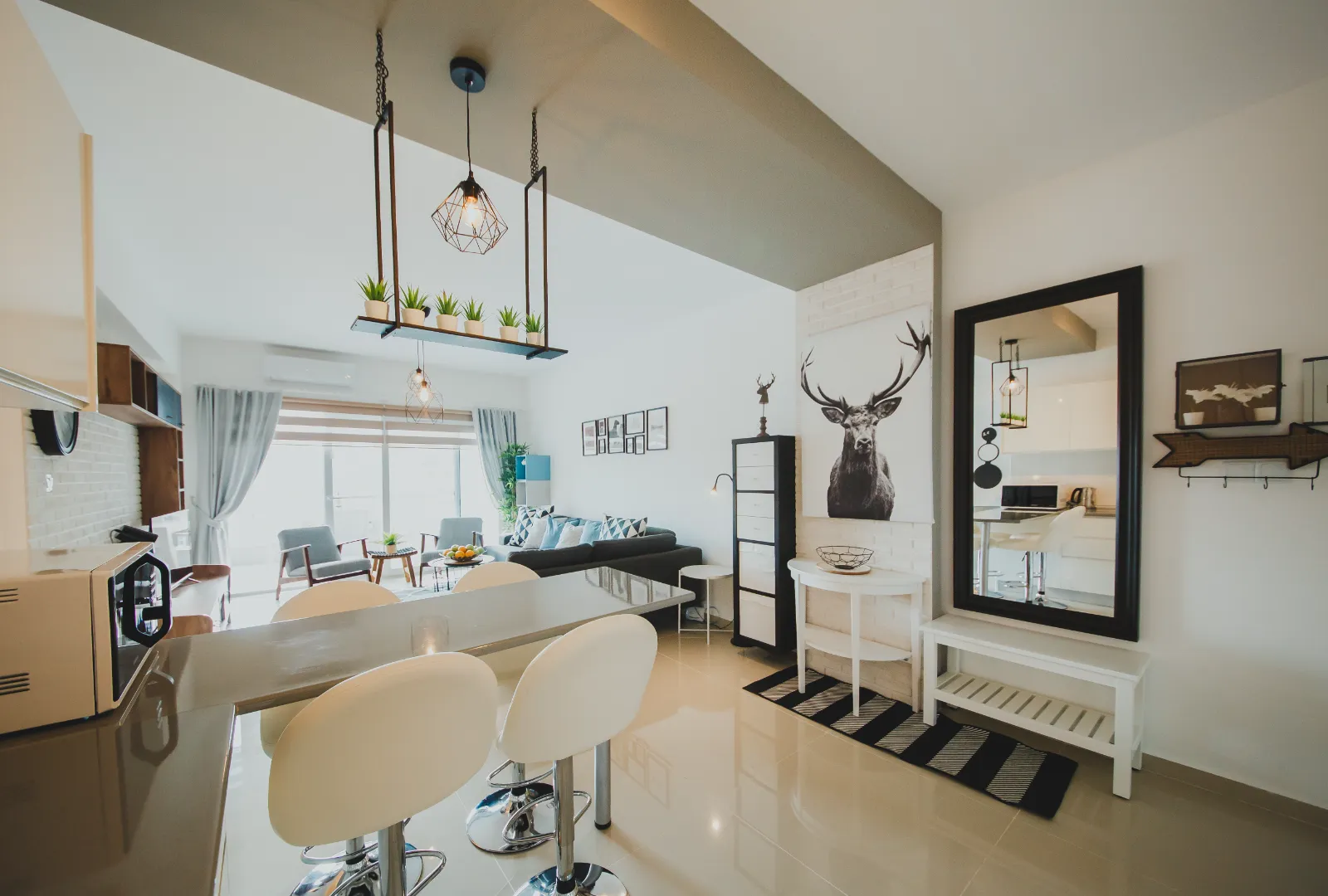 A modern, open-plan luxury apartment interior at Caesar Resort in Iskele, North Cyprus. The stylish living space features a breakfast bar with white swivel stools, a unique industrial hanging light with small potted plants, and a living area with a dark grey sofa. Nordic-inspired decor includes a large stag art piece, a minimalist black-framed mirror, and a black-and-white striped rug on polished cream tile floors.