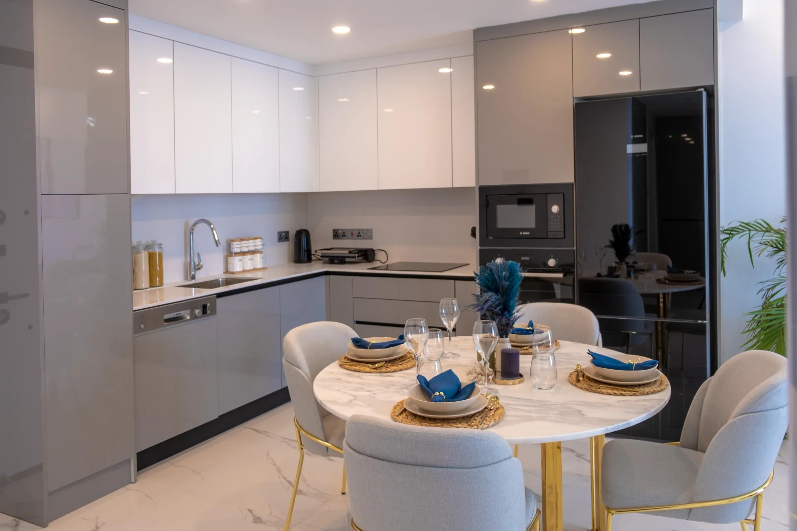 A modern luxury apartment kitchen and dining area in Kyrenia (Girne), North Cyprus. The high-gloss two-tone cabinetry in white and grey features integrated Bosch appliances, a built-in microwave, and a black glass refrigerator. A circular white marble dining table with gold legs is elegantly set for four with blue accents and plush grey chairs, reflecting the high-end interior design found in Kyrenia's newest residential developments.