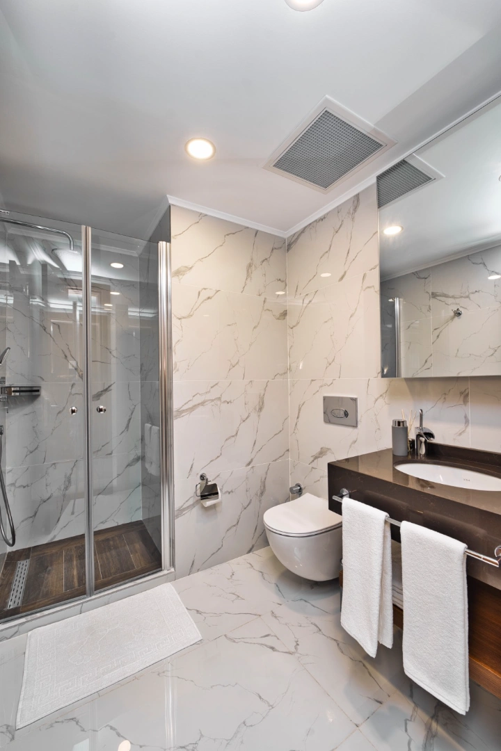 A luxurious, modern bathroom interior in Iskele, North Cyprus. The space features high-gloss Calacatta marble tiles on the walls and floors, a glass-enclosed walk-in shower with dark wood-effect flooring, and a sleek wall-mounted toilet. A dark granite vanity with a white vessel sink is adorned with fresh white towels.