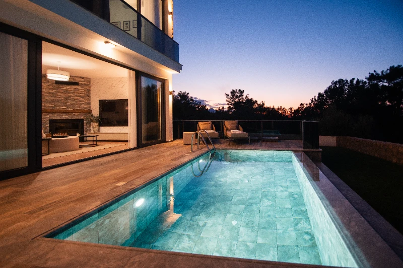 A twilight view of a luxury villa in North Cyprus featuring a private illuminated swimming pool and a large outdoor deck. The open sliding glass doors reveal a modern living room with a stone fireplace, set against a sunset sky and a silhouette of pine trees.