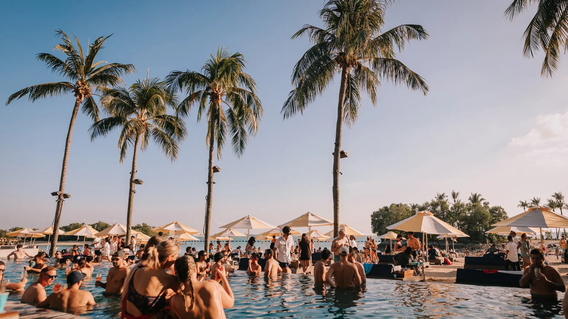 A high-energy afternoon at Pera Mackenzie Beach Club in Iskele, North Cyprus. The scene shows a crowded outdoor infinity pool filled with people socializing, surrounded by tall palm trees and cream-colored beach umbrellas. The sandy Long Beach and the Mediterranean Sea are visible in the background under a clear sky, capturing the luxurious resort lifestyle of the region.