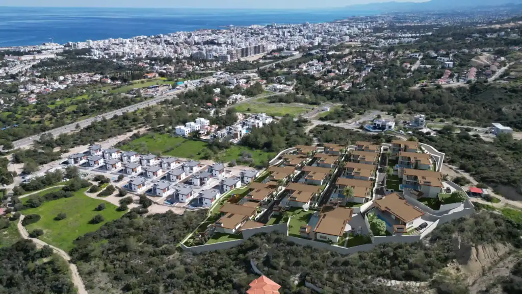 Aerial drone view of a new hillside residential development featuring modern villas with brown roofs overlooking a coastal city and the blue sea.