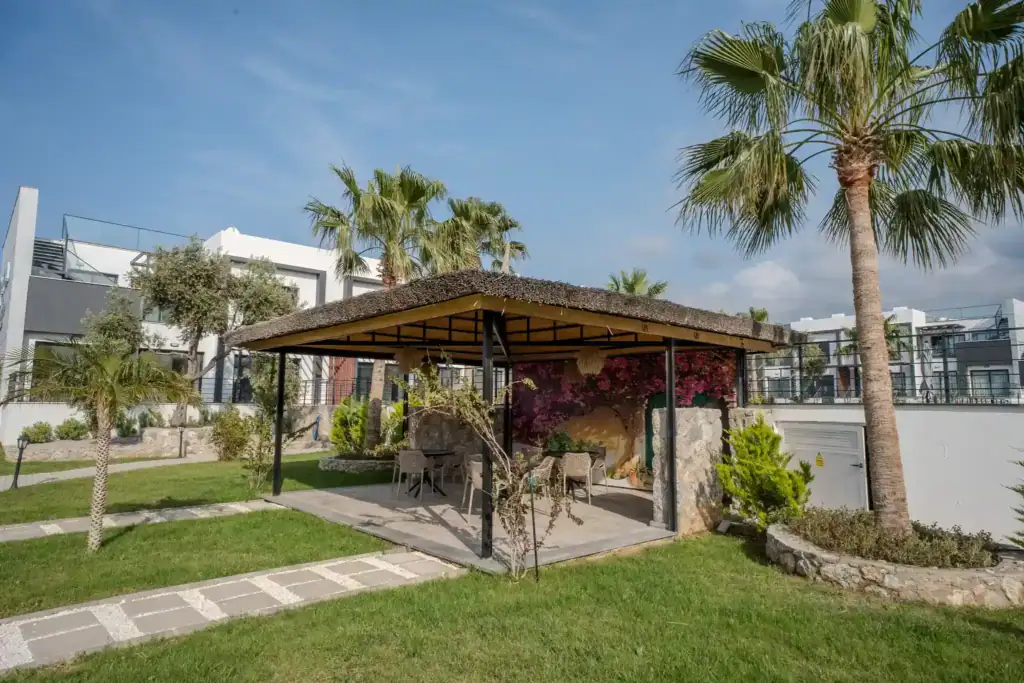 Outdoor lounge with palm trees, seating, and shaded structure in a landscaped garden.