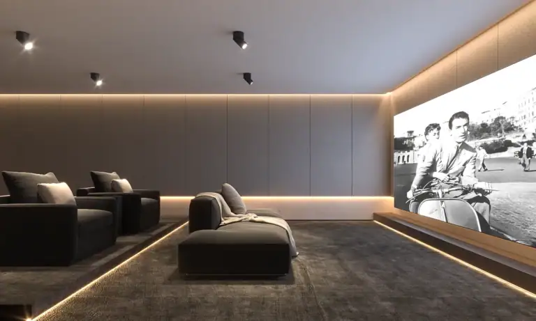 Modern luxury home theater room with dark gray tiered seating, ambient LED accent lighting, and a large projection screen showing a black-and-white movie.