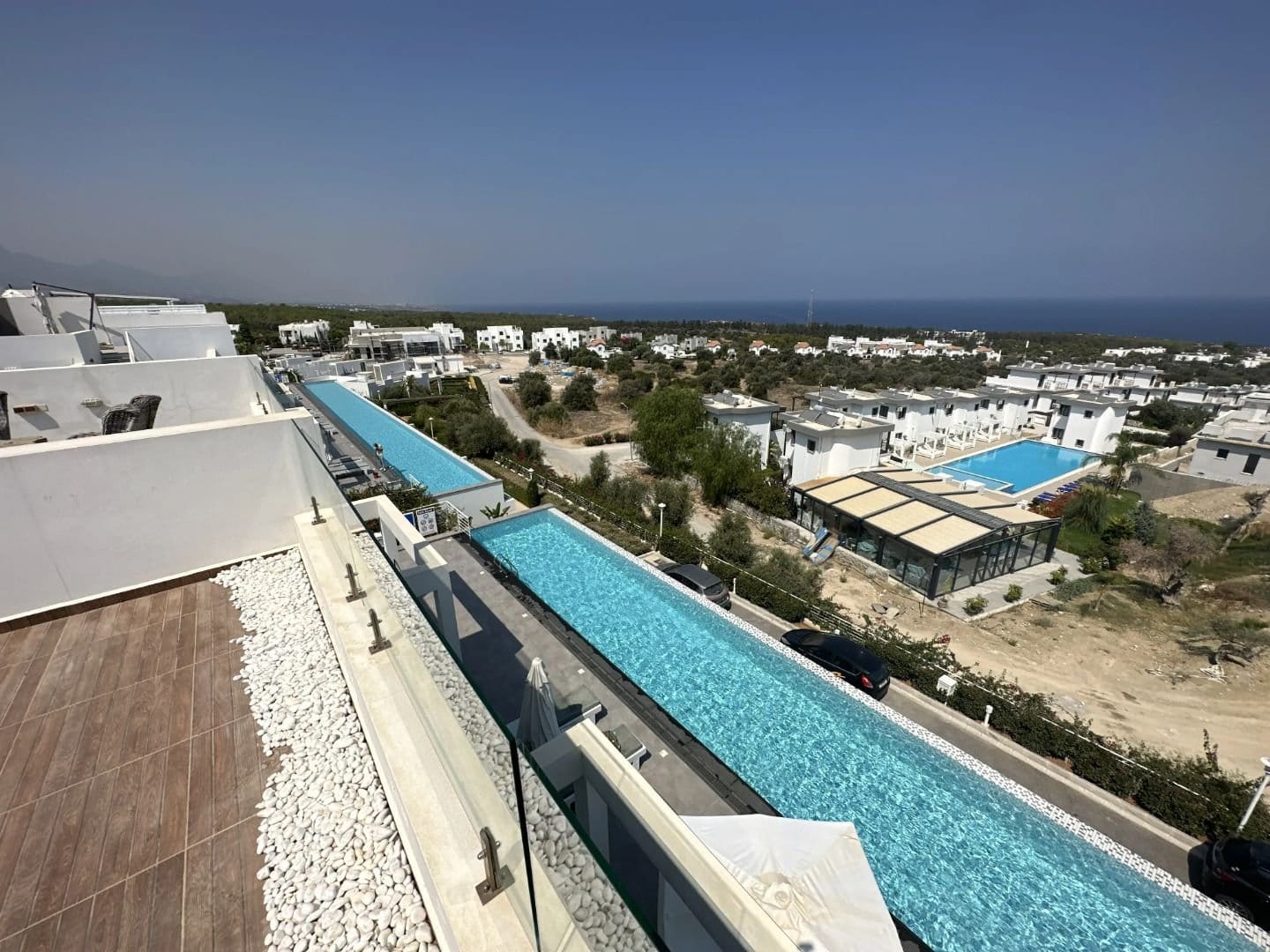 A lush infinity pool overlooking North Cyprus coastline and residential area, with modern white buildings and clear blue sky.